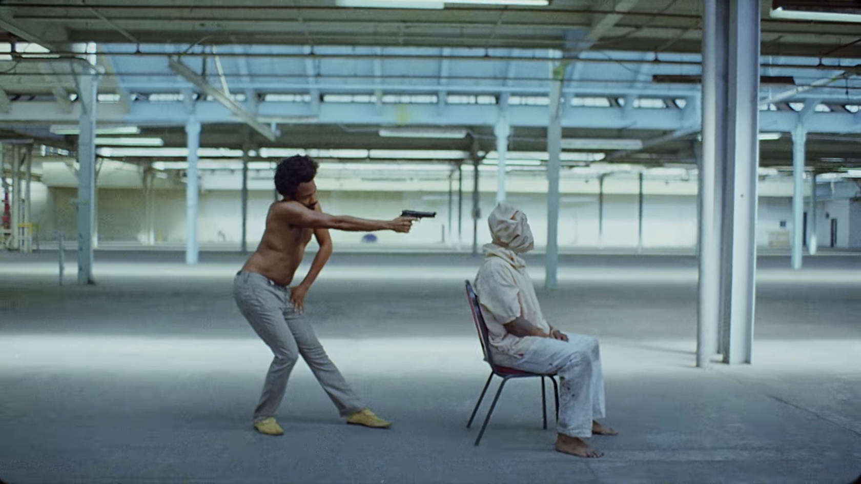 A still from the video "This Is America" showing Don Glover/Childish Gambino pointing the gun at a man sitting on a chair, wearing a hood over his head