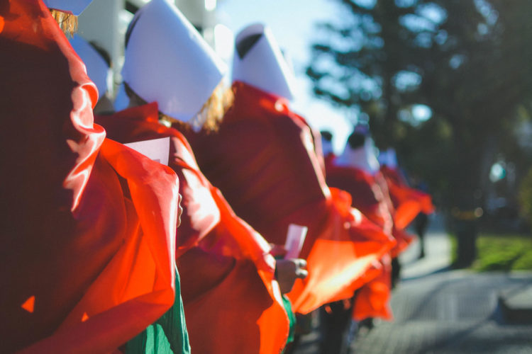 Women in a line, dressed like characters from The Handmaid's Tale (dressed in red with a white veil on the head)